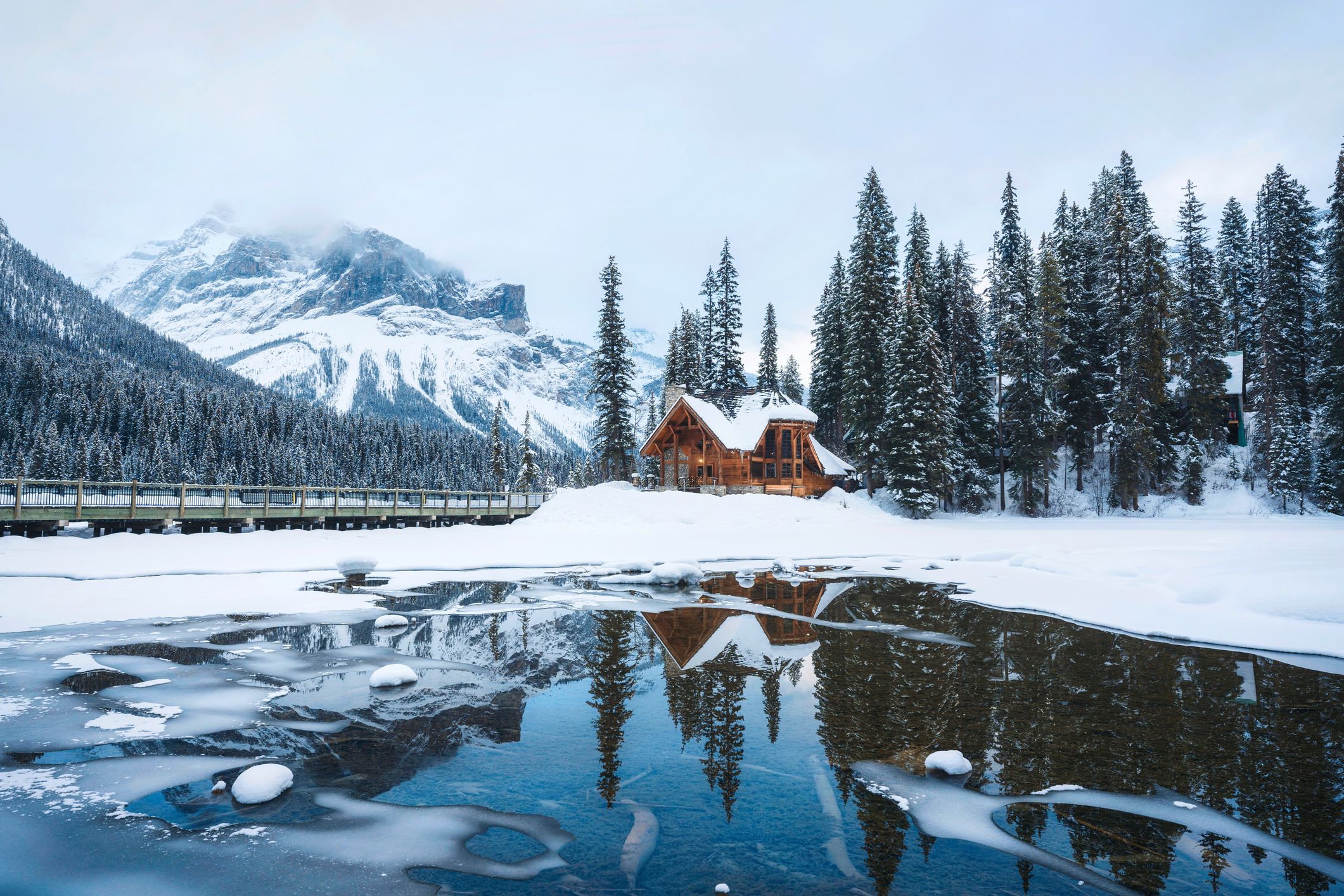 cottage house in a winter snowing day with mountain, trees, and by the water
